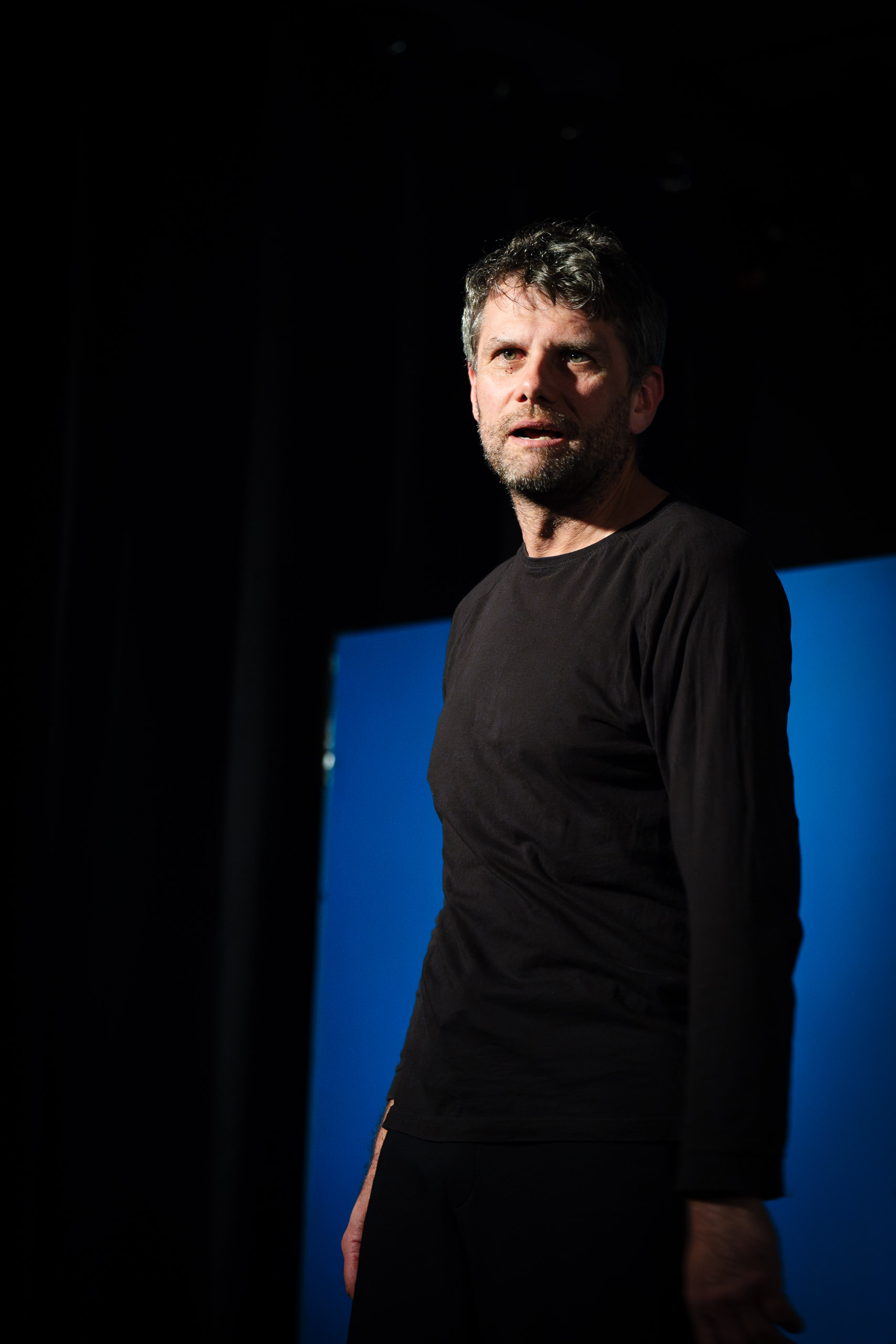A man in a black shirt stands against a blue backdrop, looking o