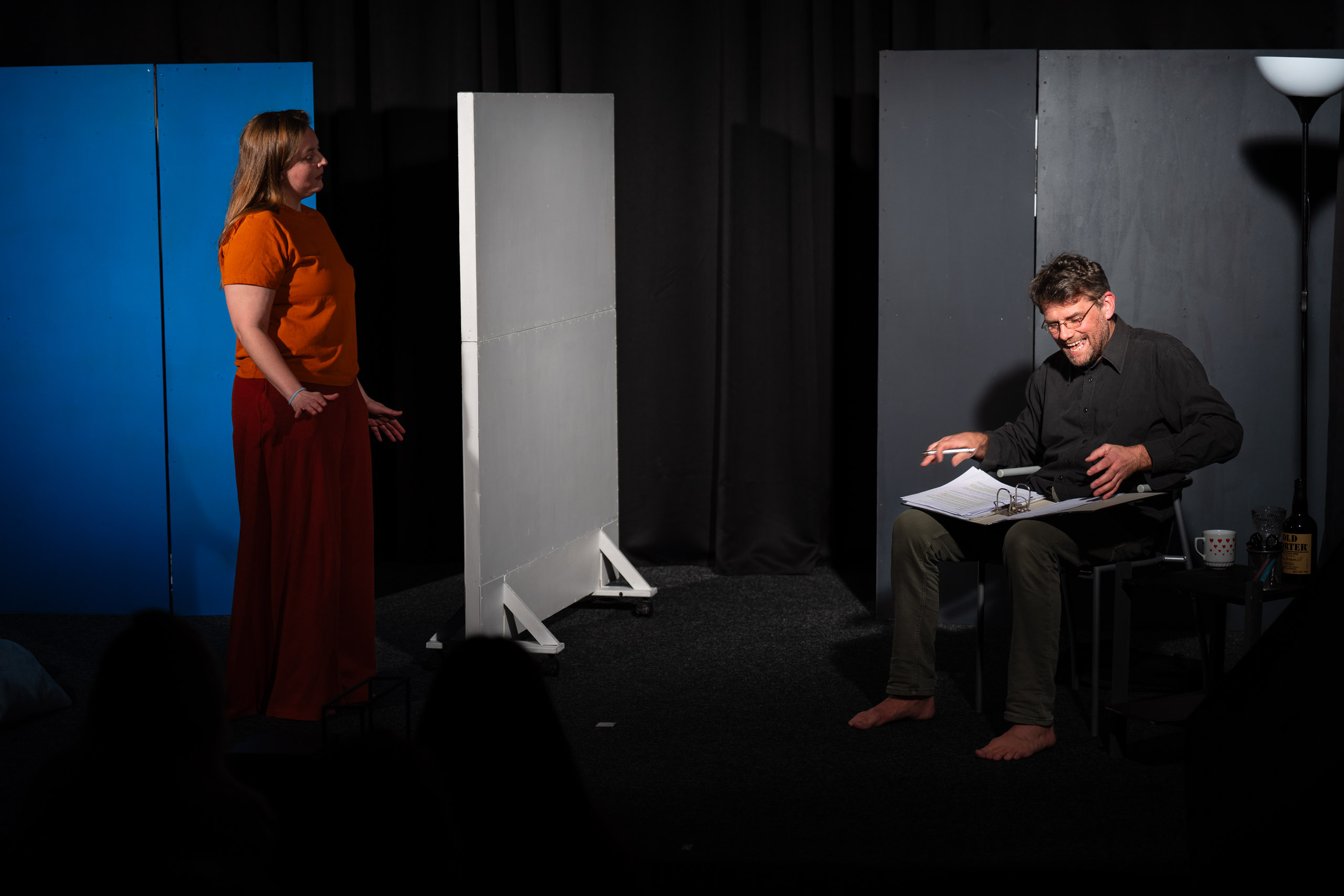 A woman stands and gestures towards a man sitting with papers, p