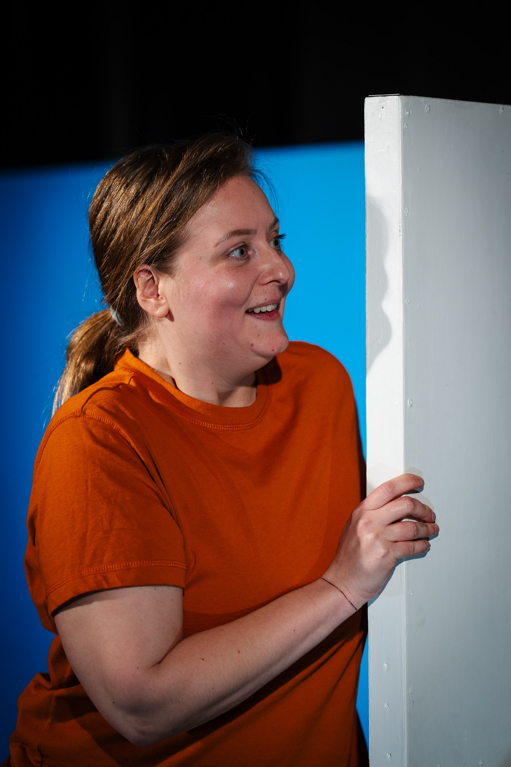 A woman in an orange shirt peeks around a white corner with a bl