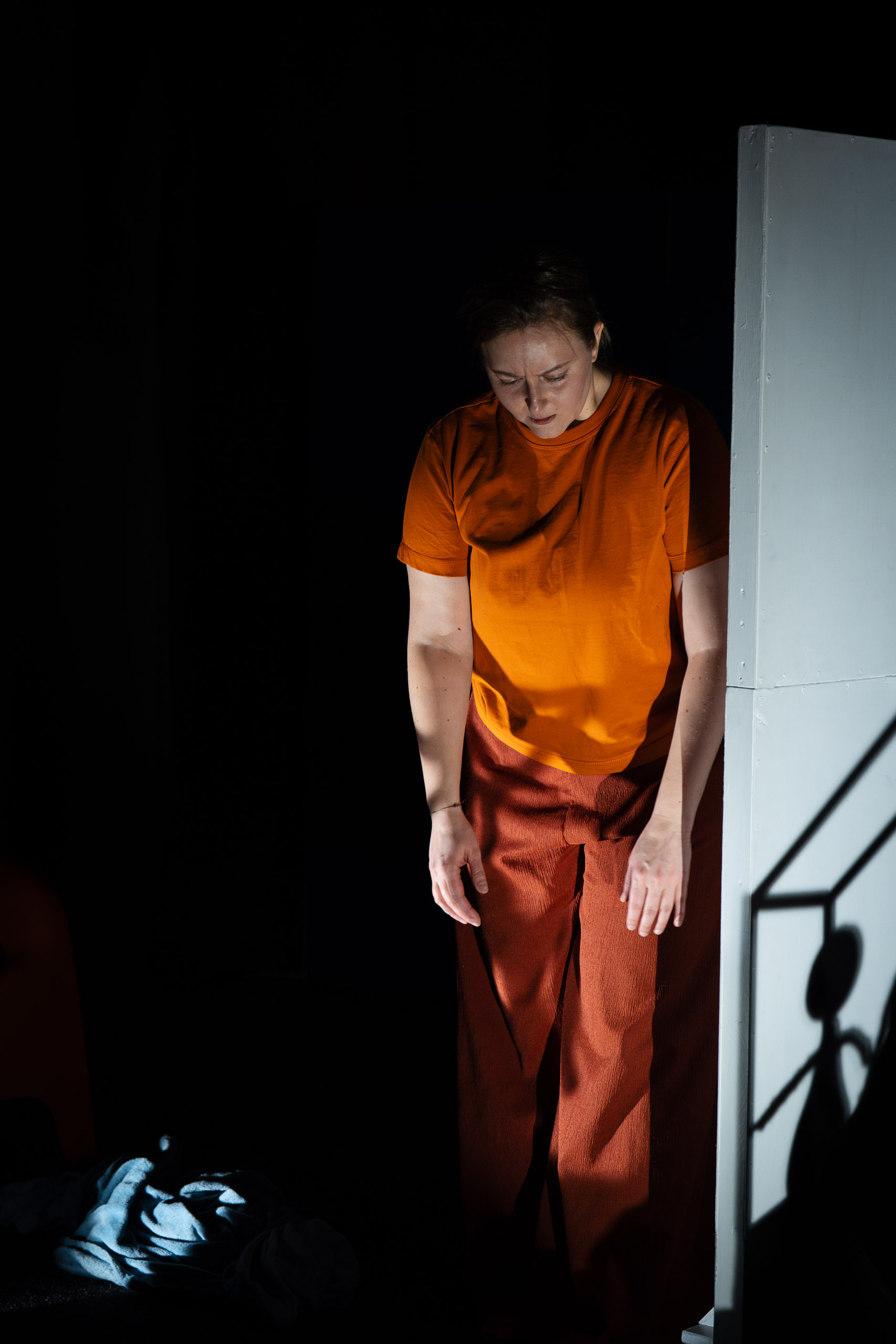 A woman in an orange shirt and brown pants stands in dim light,