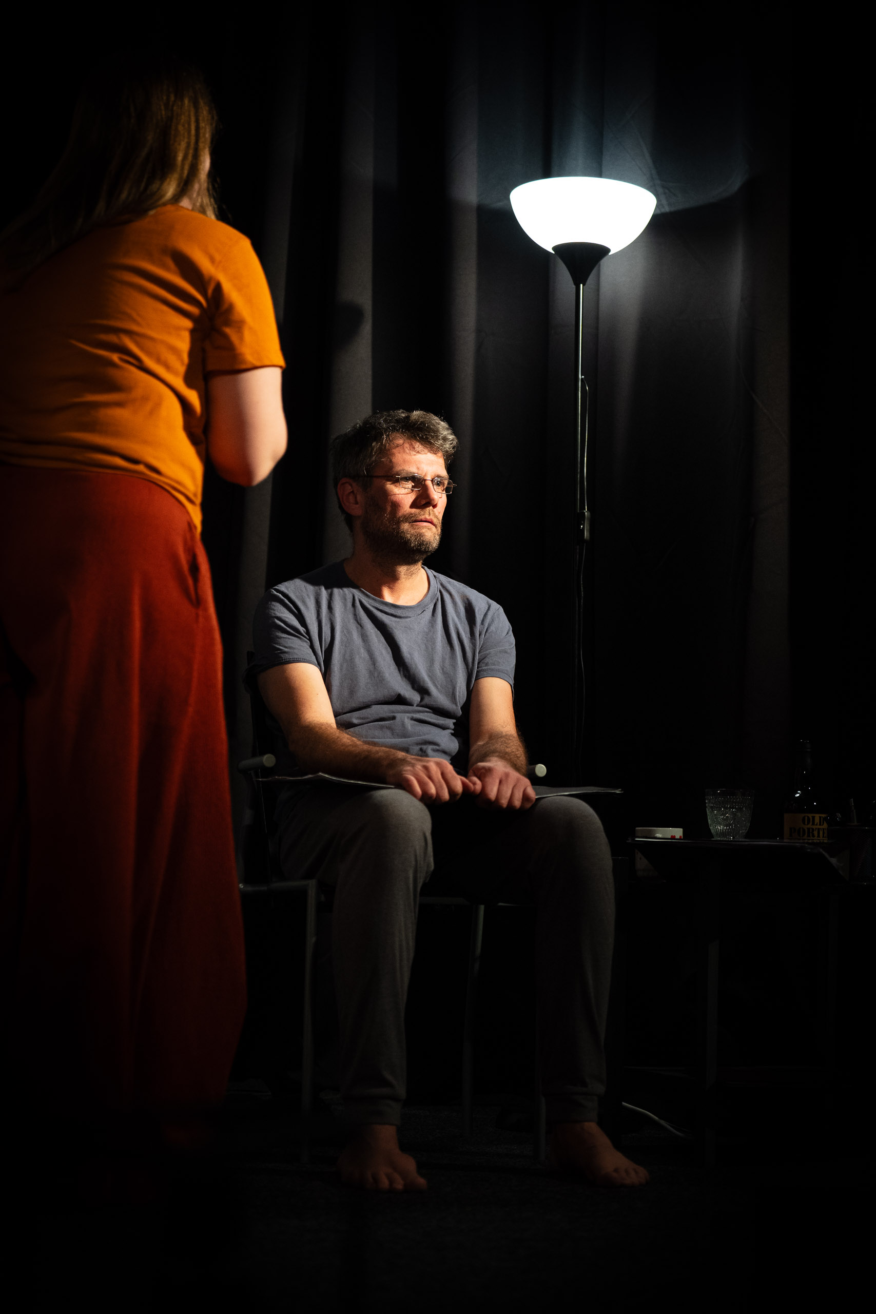 A man sits in a chair under a lamp while a woman stands nearby.