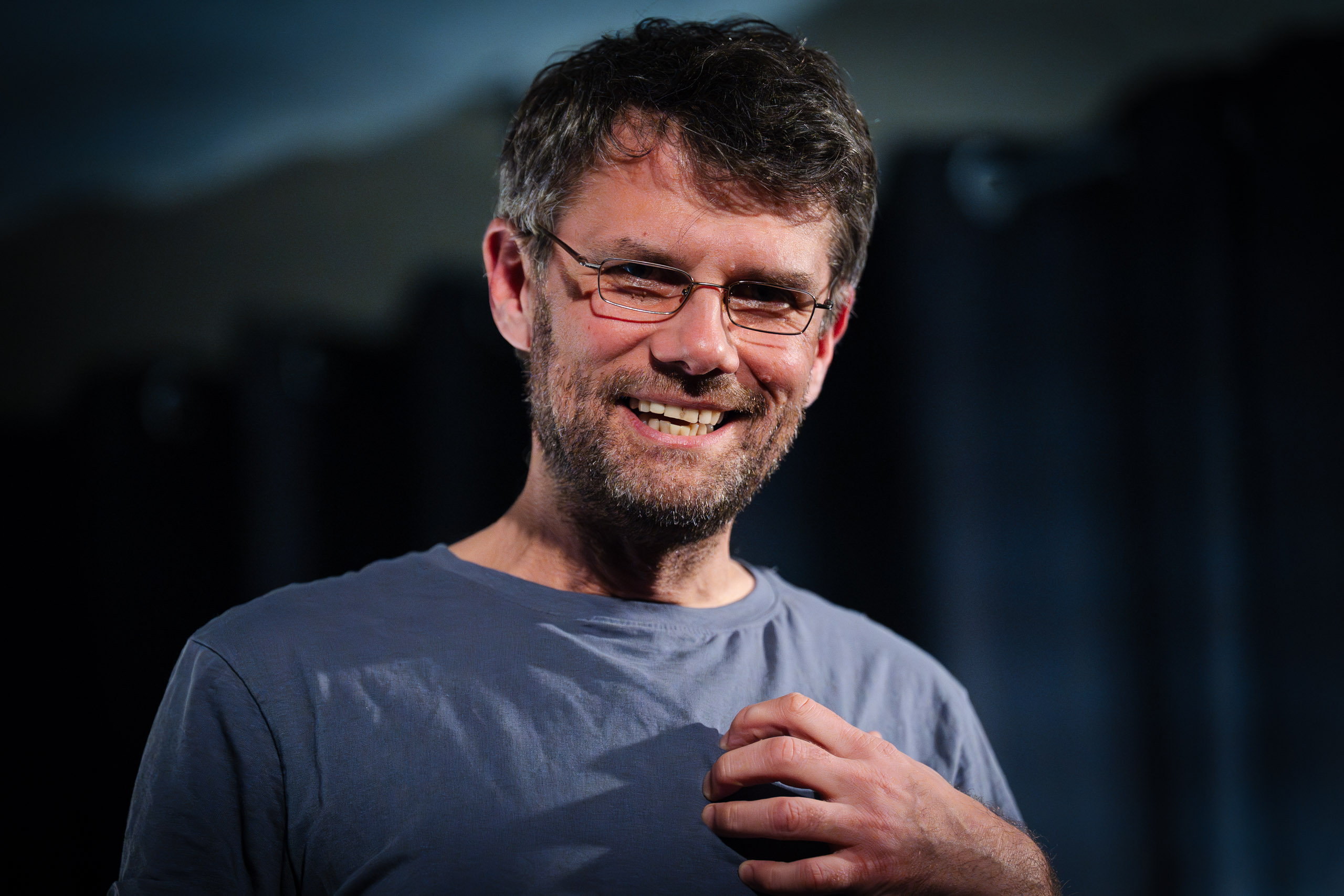 A man with glasses and a beard smiles and touches his chest.