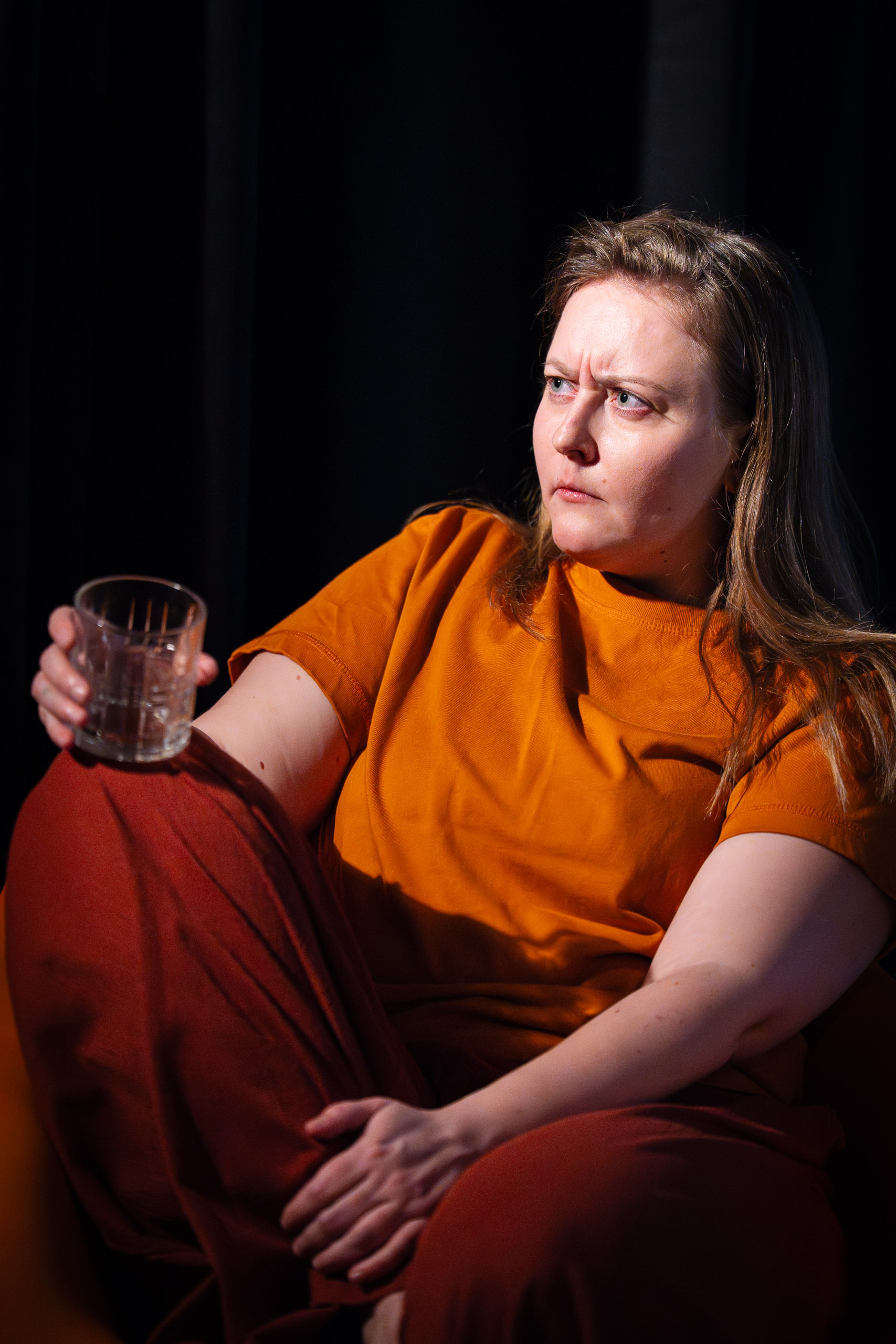 A woman with an orange shirt and red pants sits holding a glass