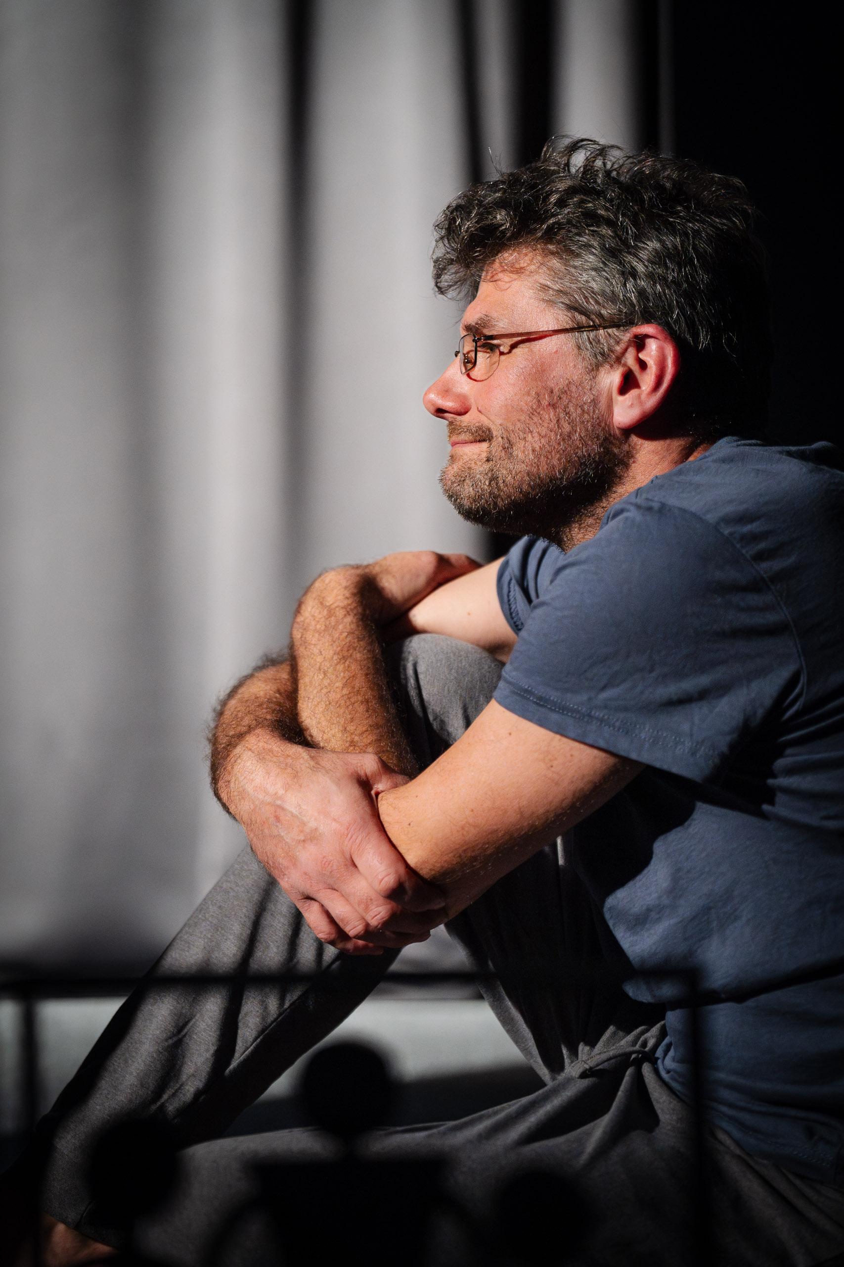 A man with glasses sits with his arms crossed, looking away from