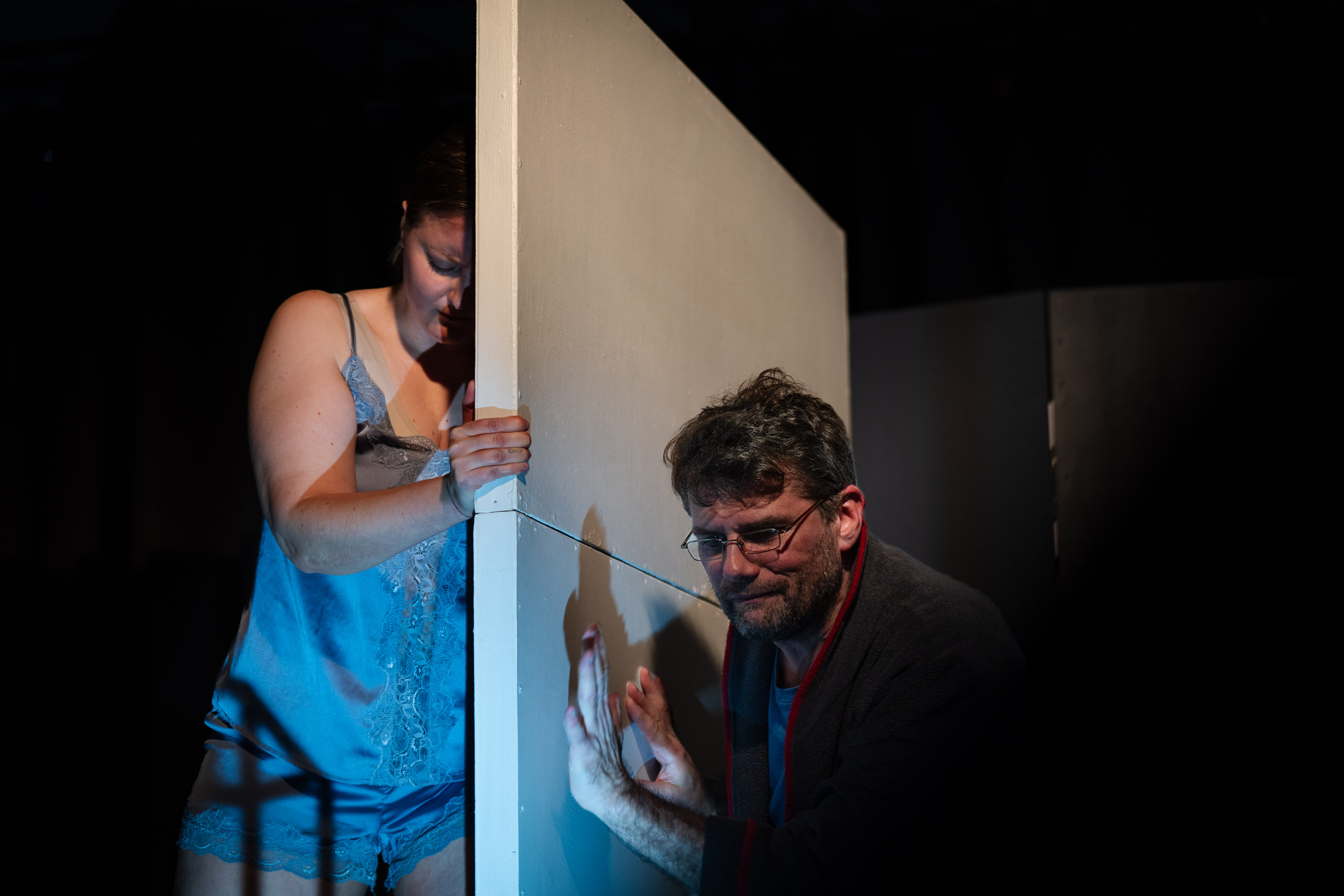 A woman in blue lingerie peeks around a corner as a man in glass