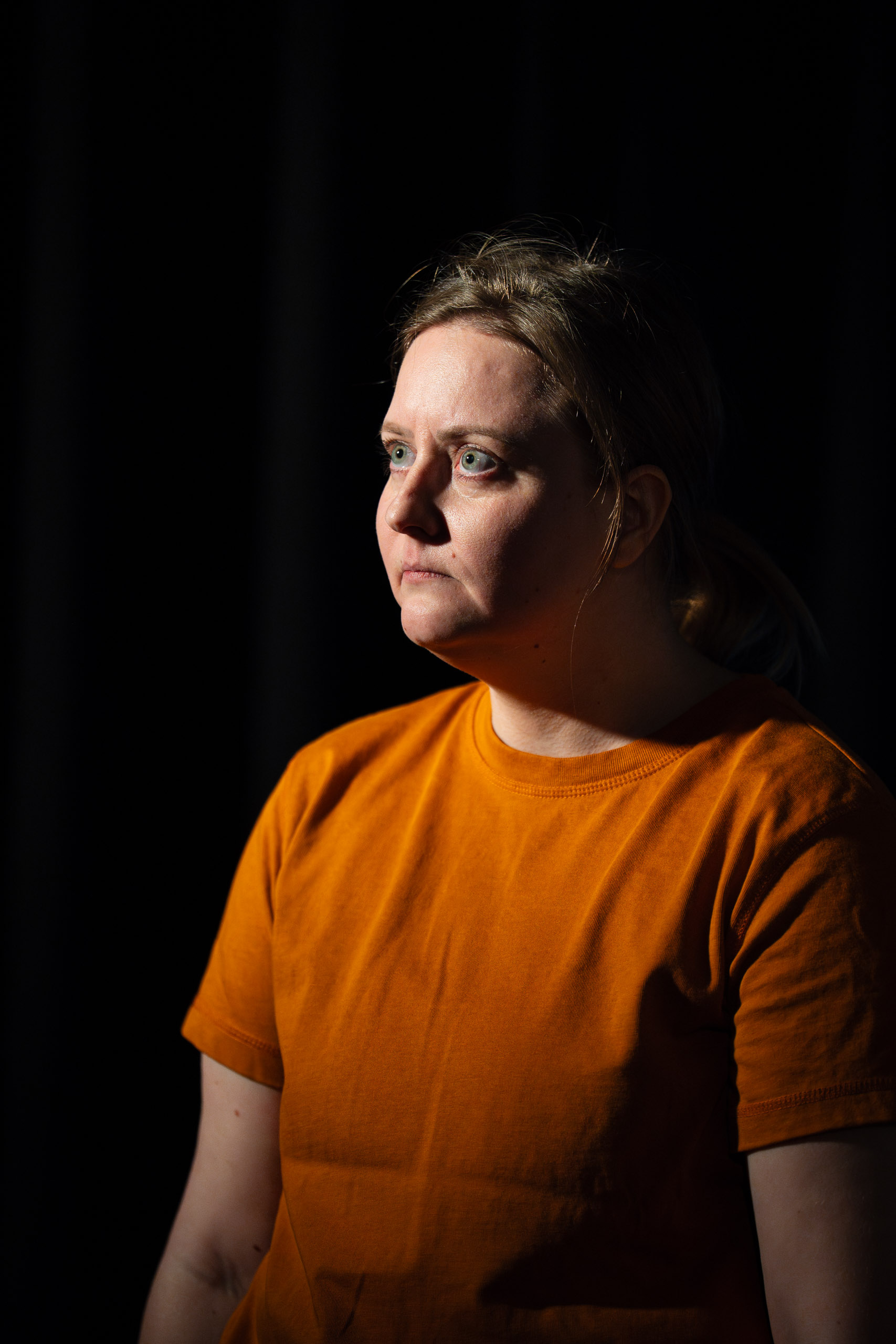 A woman in an orange t-shirt looks off to the side with a seriou