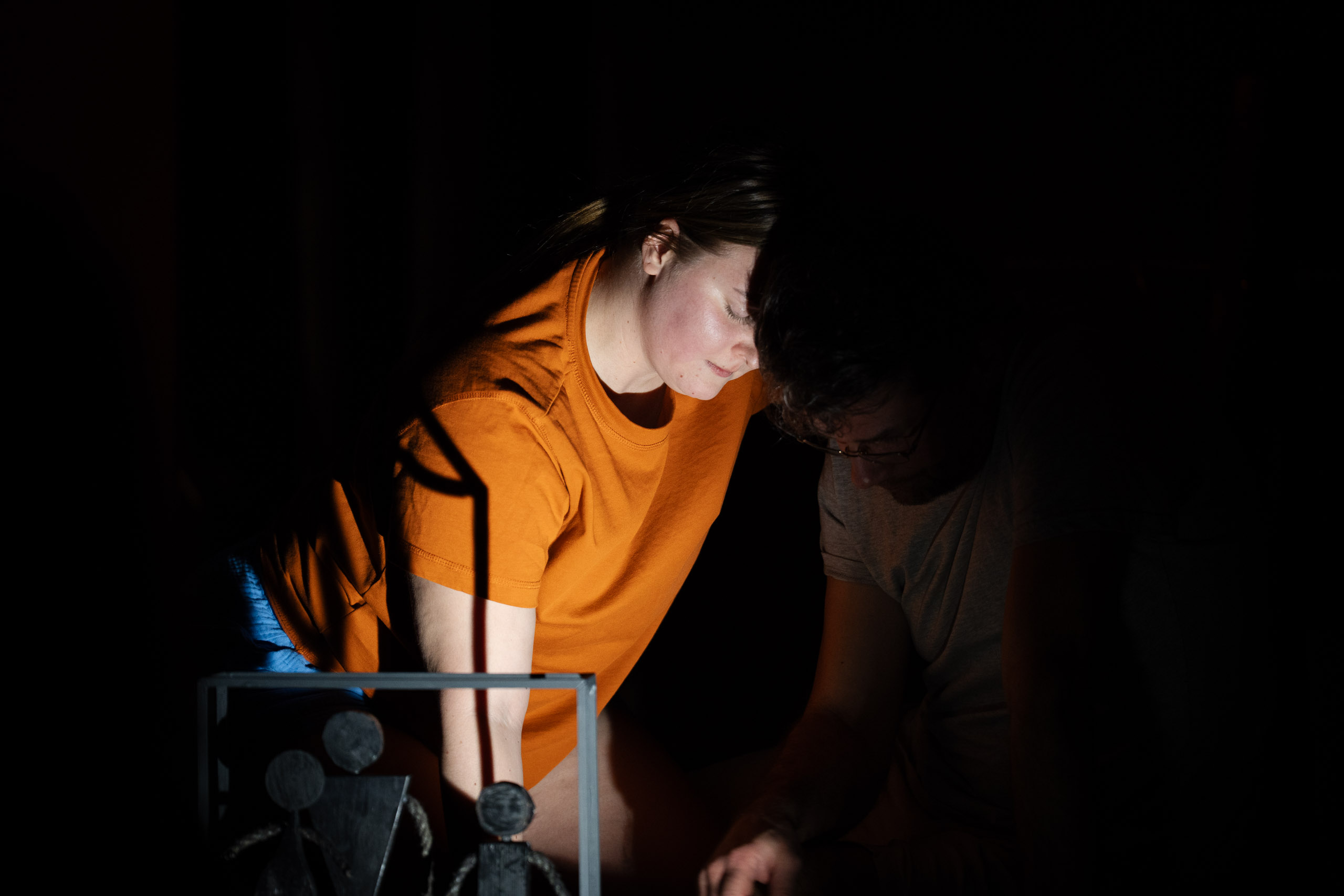 Two people in the dark, one wearing an orange shirt, lean in tog