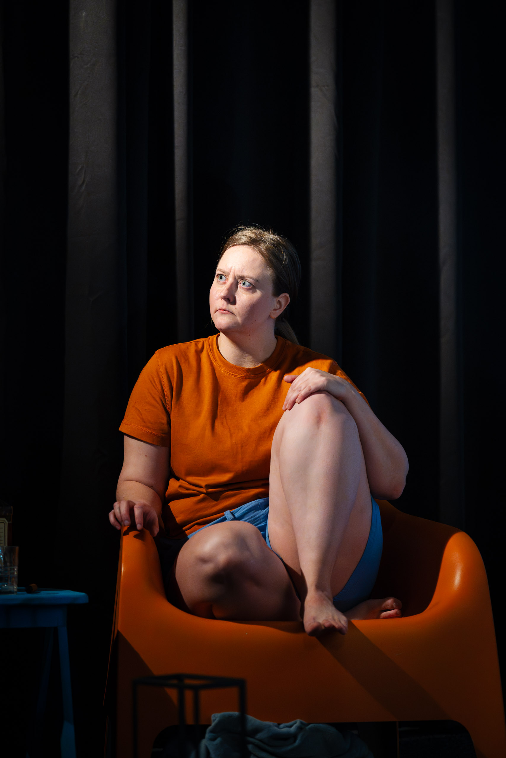A woman sits in an orange chair, looking away with a pensive exp