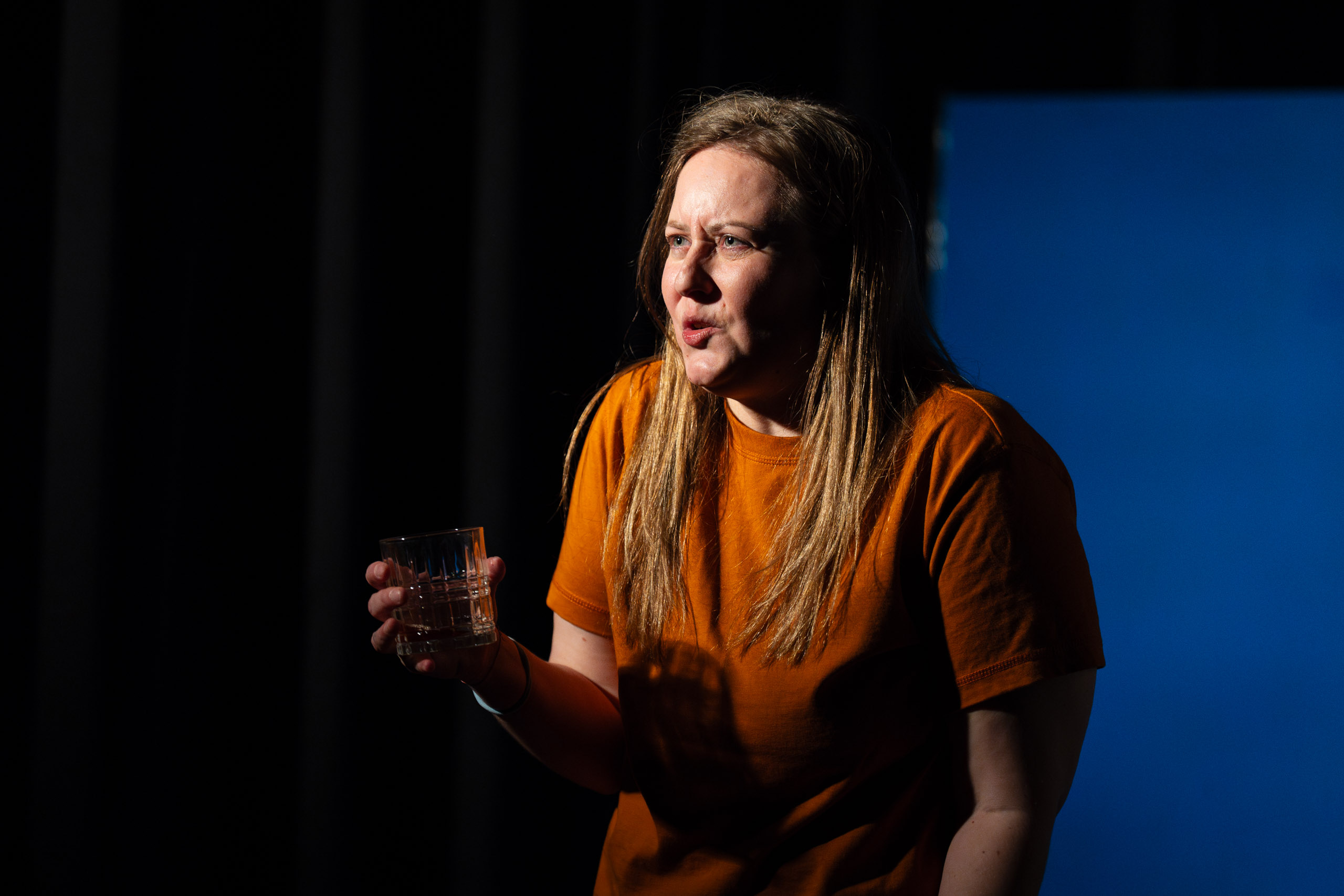 A woman in an orange shirt holds a glass and speaks with a drama