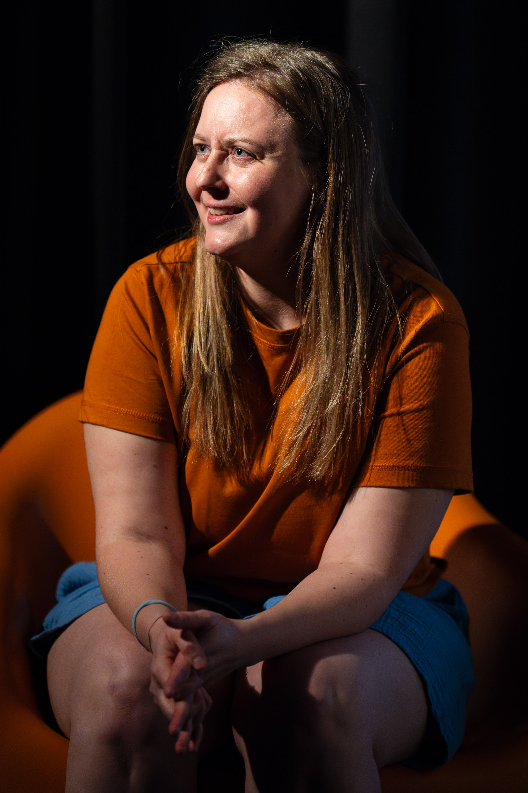 A woman in an orange t-shirt and blue shorts smiles while sittin