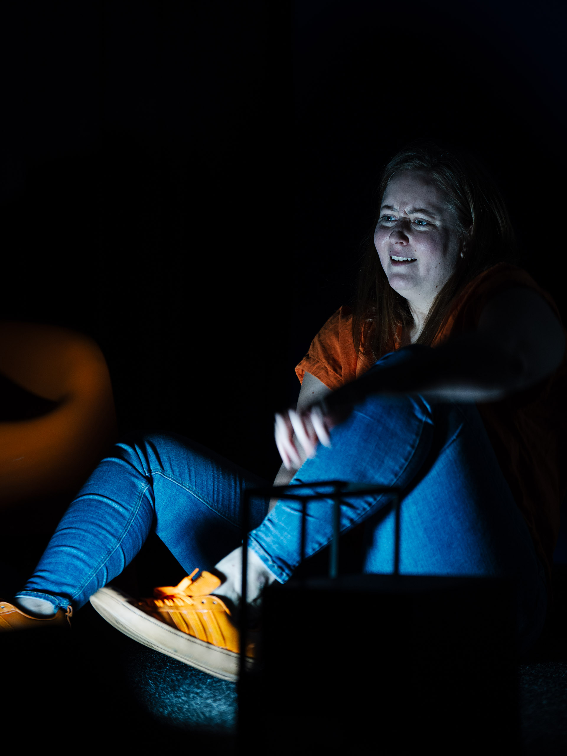 A woman sits in the dark, looking distressed and scared at somet